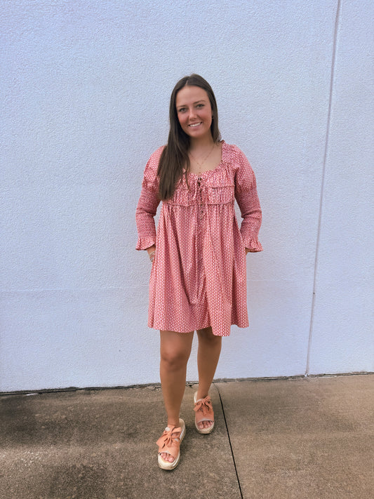 Model wearing a pink and rust geometric print babydoll dress featuring a smocked back, adjustable tie-back detail, puff sleeves, and a flowy silhouette.