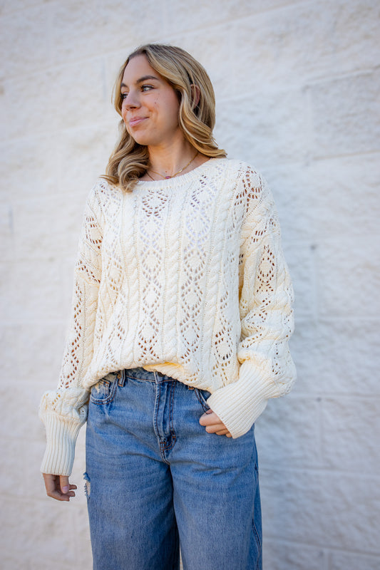 Cream cable knit sweater featuring intricate open-knit detailing, a relaxed fit, ribbed crew neckline, oversized ribbed cuffs, and long sleeves for a cozy fall and winter look.