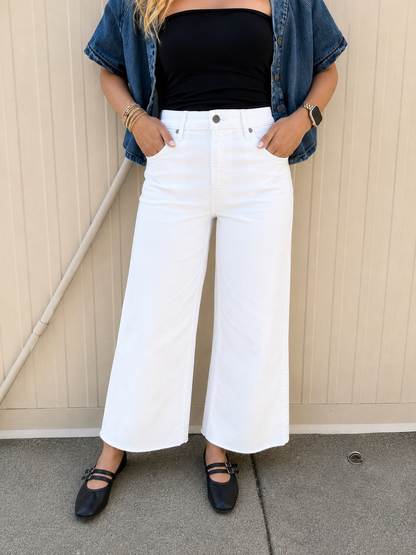 Women’s white cropped wide leg jeans styled with a black strapless tube top and medium wash cropped denim button-up shirt. Features a high-rise fit, clean white denim, cropped wide leg silhouette, raw hem, and side slit detail, paired with black Mary Jane flats for a casual summer outfit.