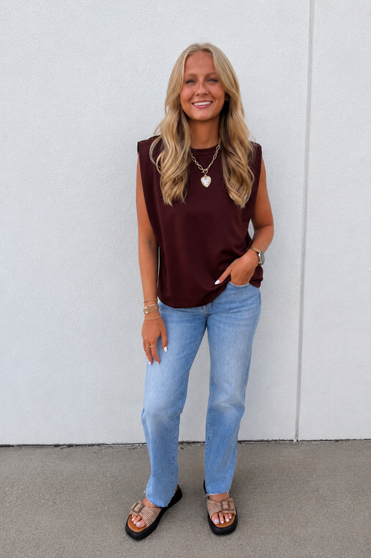 A woman stands against a light gray wall wearing a sleeveless brown top, light blue jeans, and brown slide sandals.