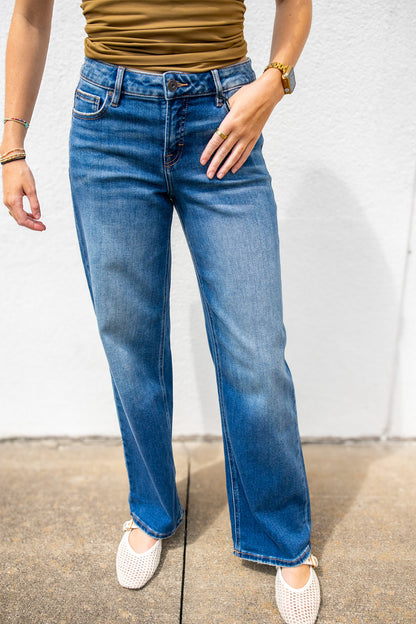 A person wearing dark-wash blue straight-leg jeans, a brown ribbed top, and white knit slip-on shoes, standing against a white wall.