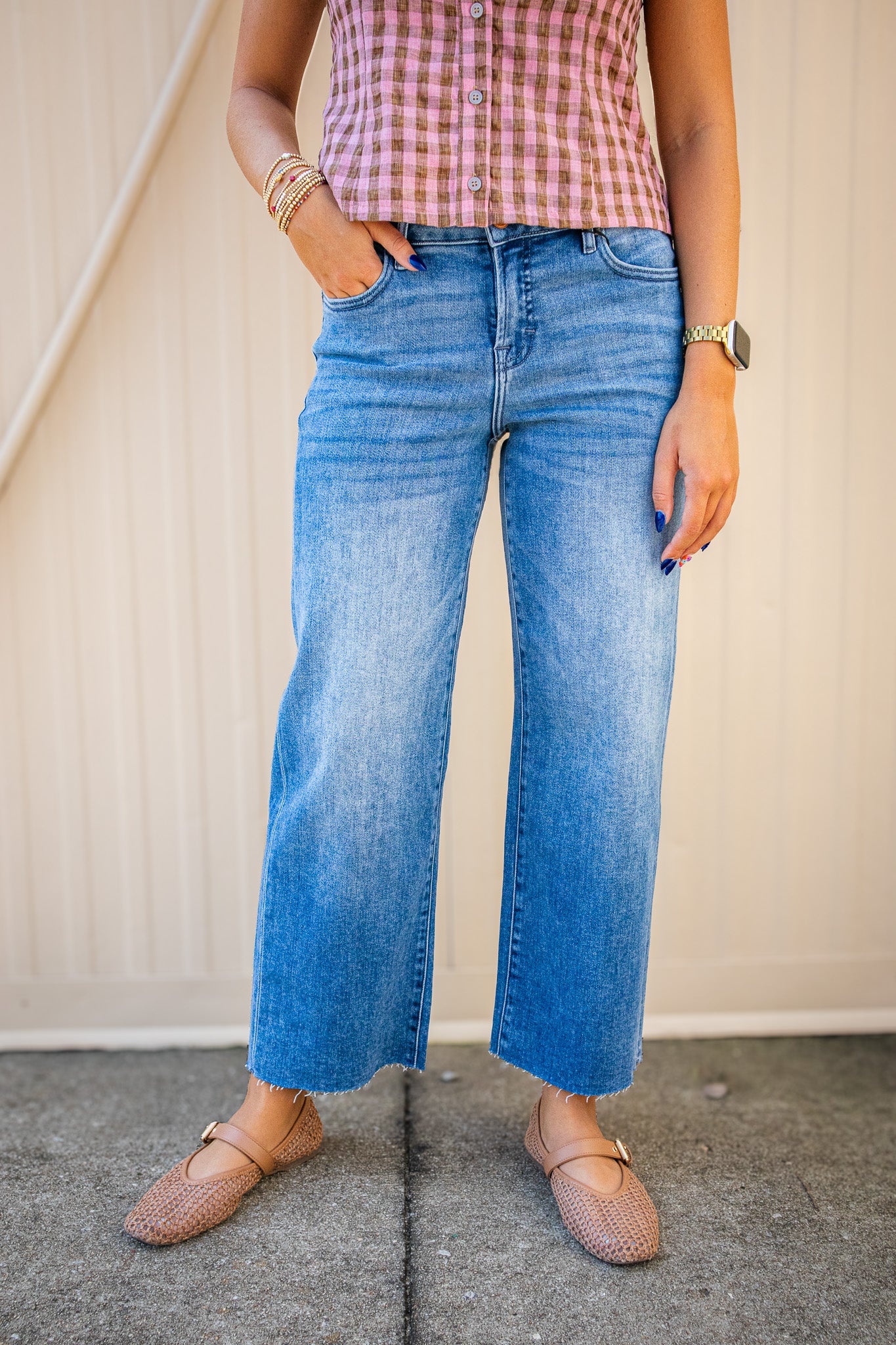 A person wearing light-wash blue wide-leg cropped jeans, a pink and white gingham top, and tan flat shoes, standing against a beige paneled wall.