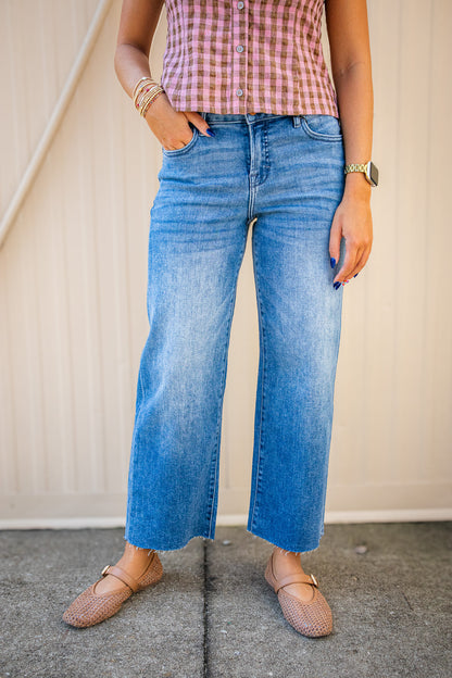 A person wearing light-wash blue wide-leg cropped jeans, a pink and white gingham top, and tan flat shoes, standing against a beige paneled wall.