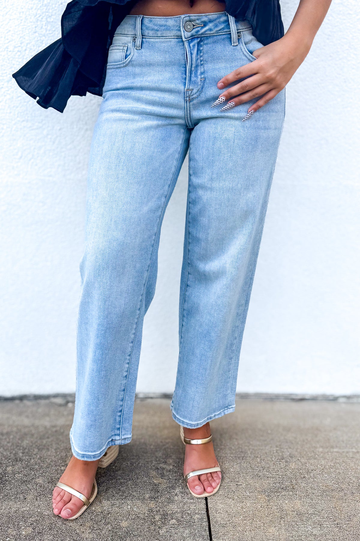 A person wearing light-wash blue wide-leg cropped jeans with a dark navy ruffled top and metallic heeled sandals, standing against a white wall.