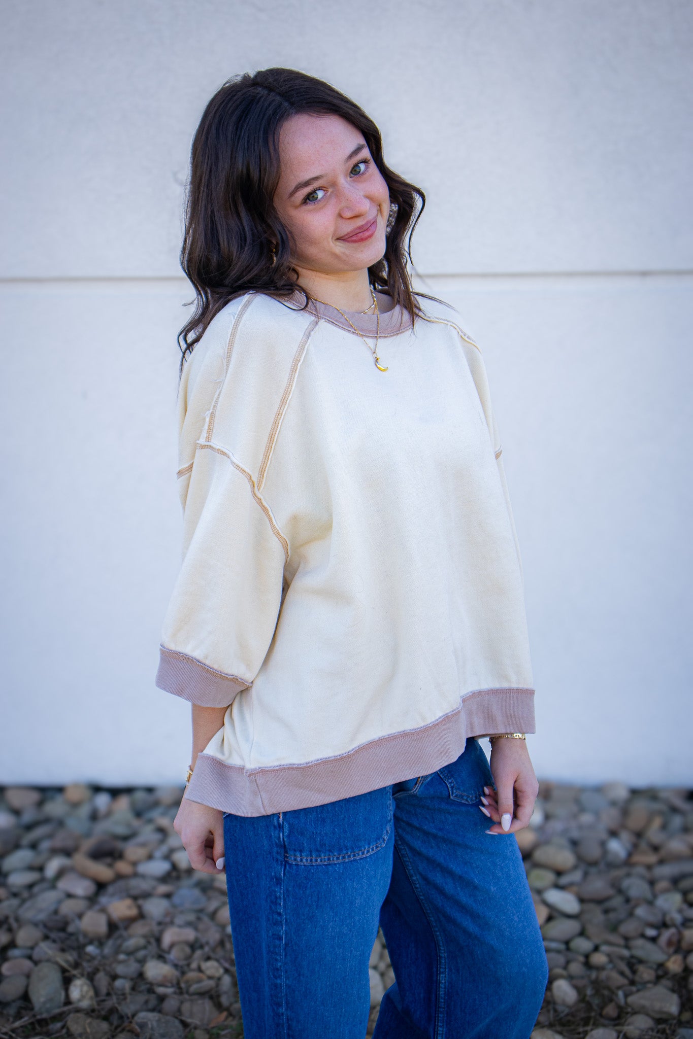 Woman wearing a light-colored sweater and blue jeans standing against a white wall.