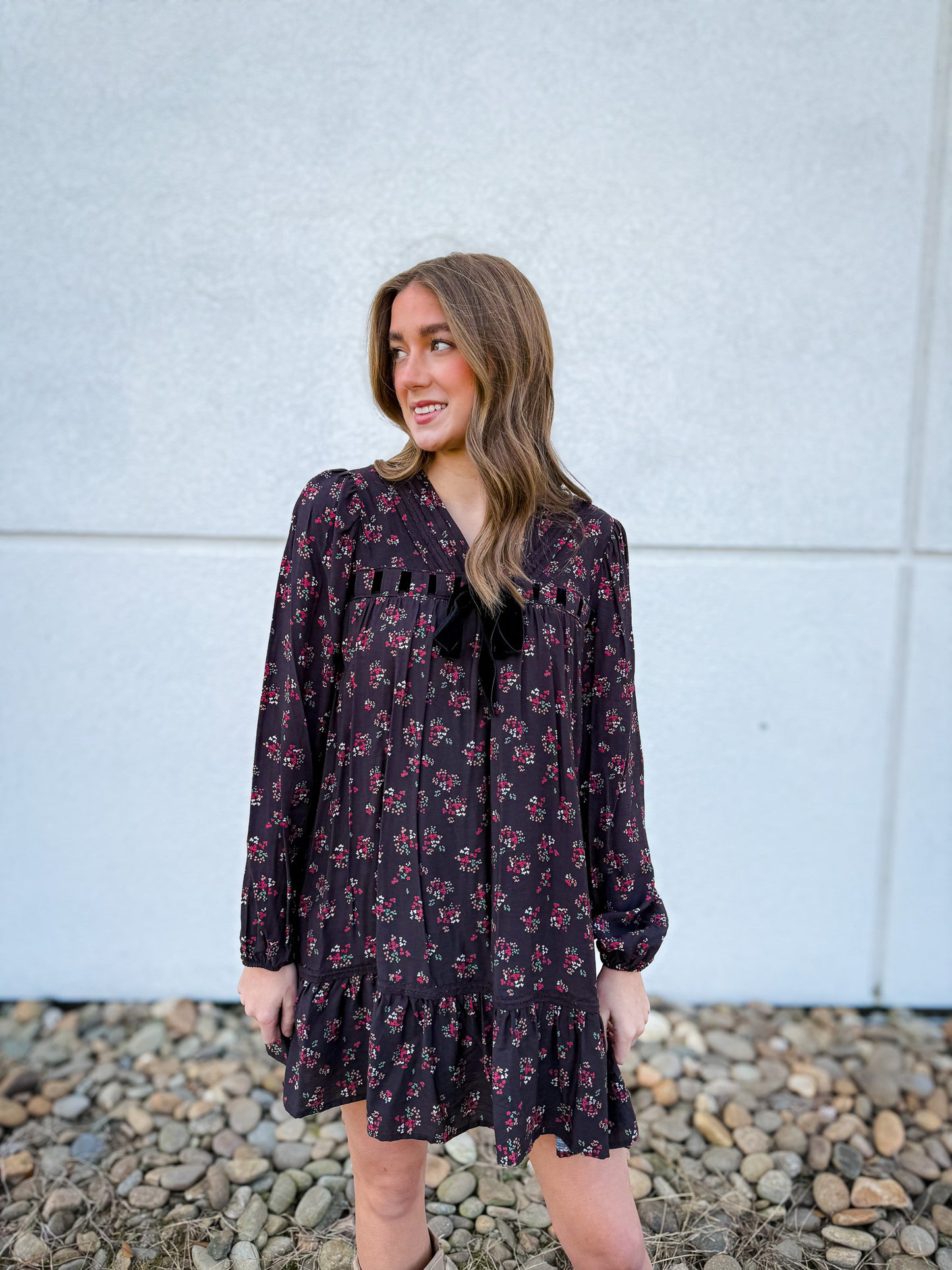 Woman wearing a black floral dress standing against a light gray wall with pebbles at the bottom.