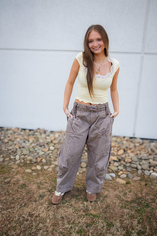 Woman wearing a light-colored top and gray pants standing outdoors with a white wall and stones in the background.