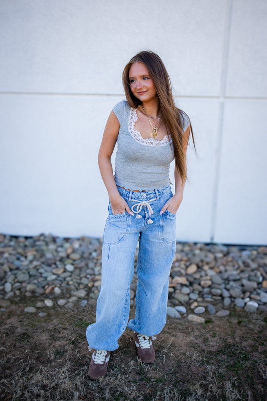 Woman wearing a light blue sleeveless top and jeans standing against a white wall with rocks on the ground.