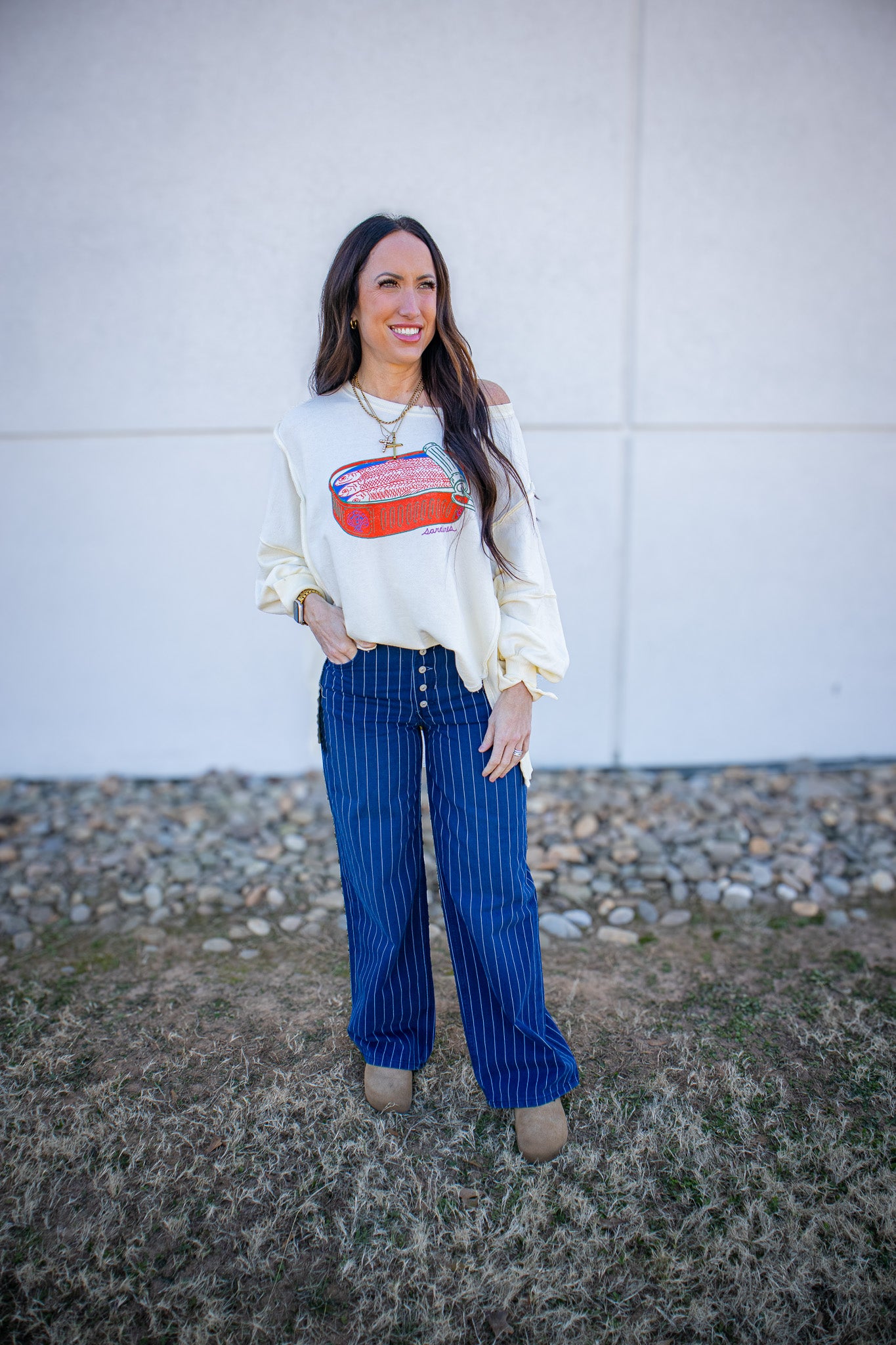 Woman wearing a white sweater with a colorful design and blue striped pants standing in front of a white wall.