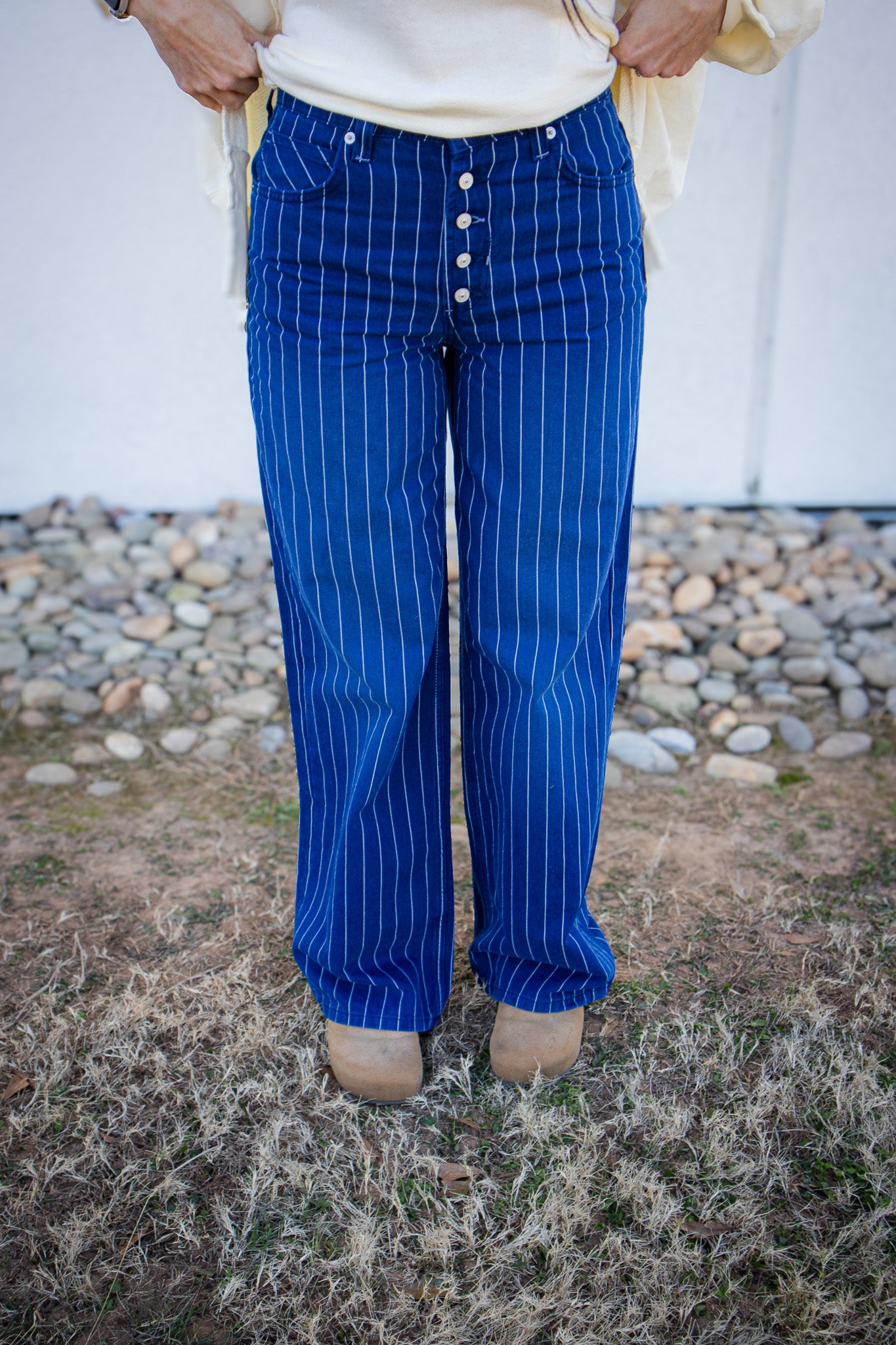 Person wearing blue pinstripe pants with a white shirt, standing on grass with a stone wall in the background.
