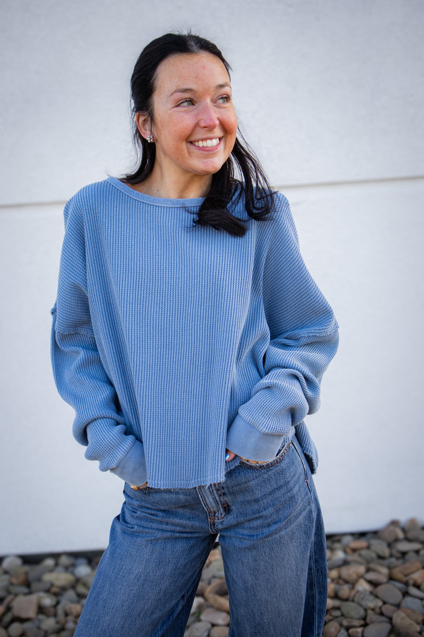 Woman wearing a blue, waffle knit top and jeans standing against a white wall.