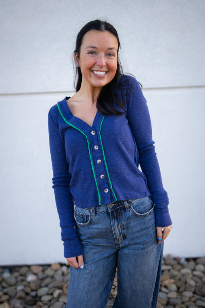 Woman wearing a blue long sleeve top with green trim and jeans standing against a white wall.