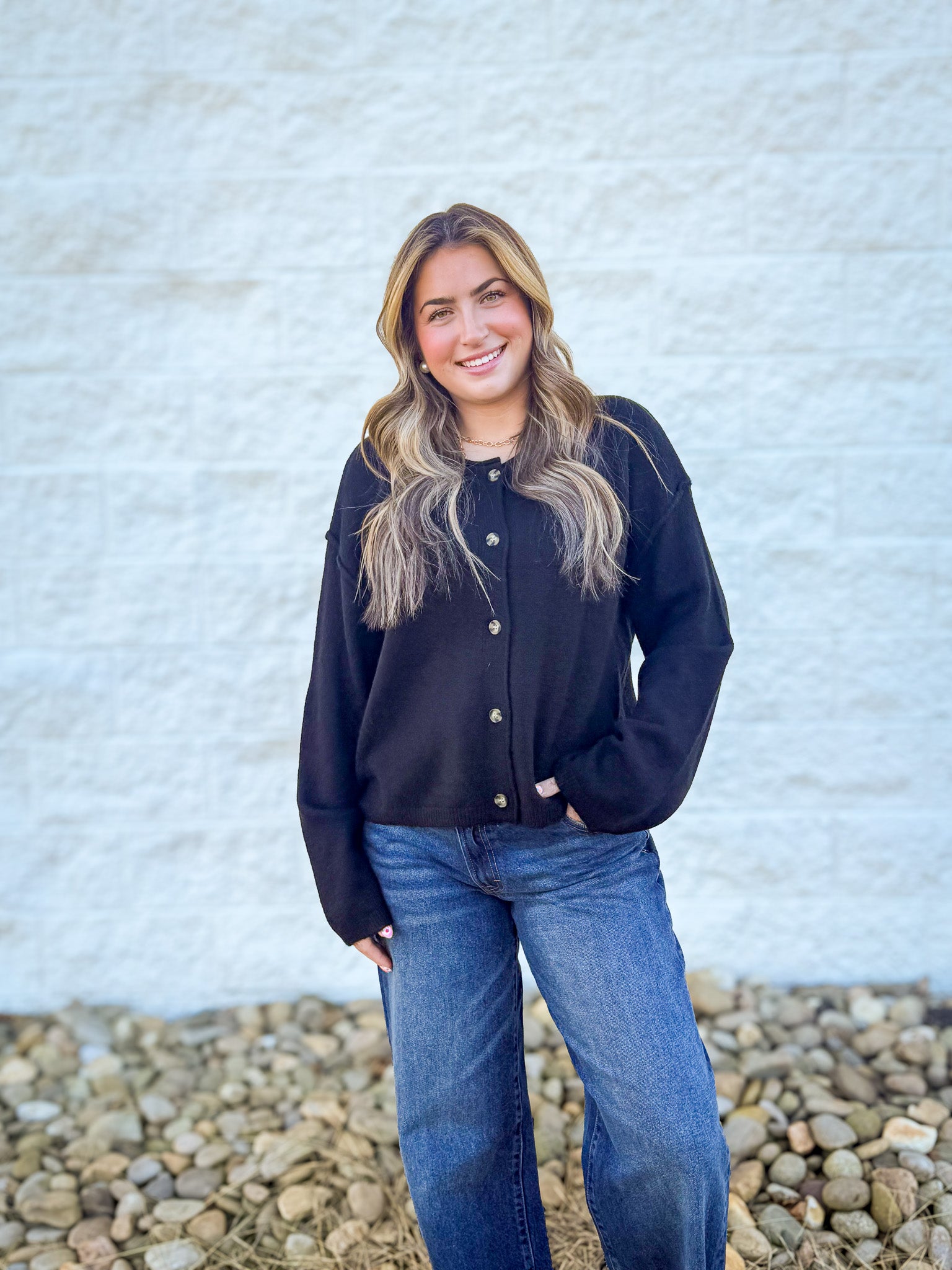 Woman wearing a black jacket and blue jeans standing against a textured white wall.