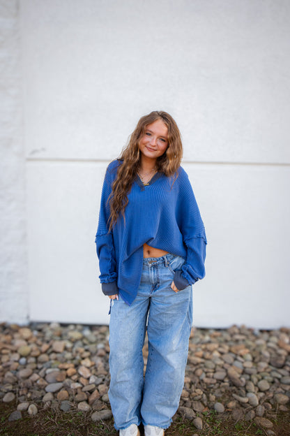 Woman wearing a blue top and jeans standing against a white wall with a stone ground.