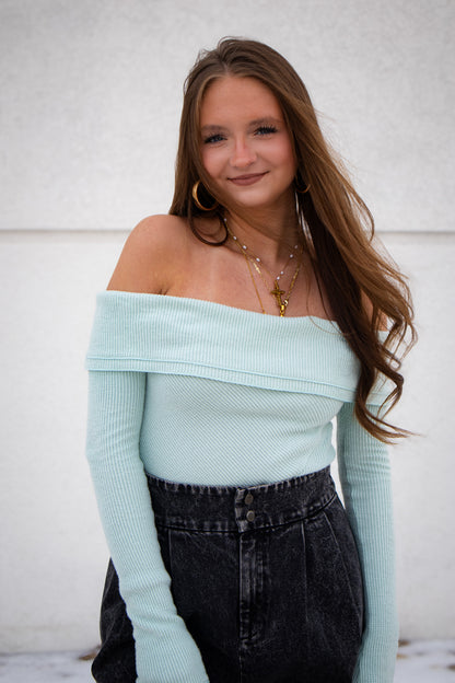 Woman wearing a light blue off-shoulder bodysuit tucked into dark jeans against a white background.
