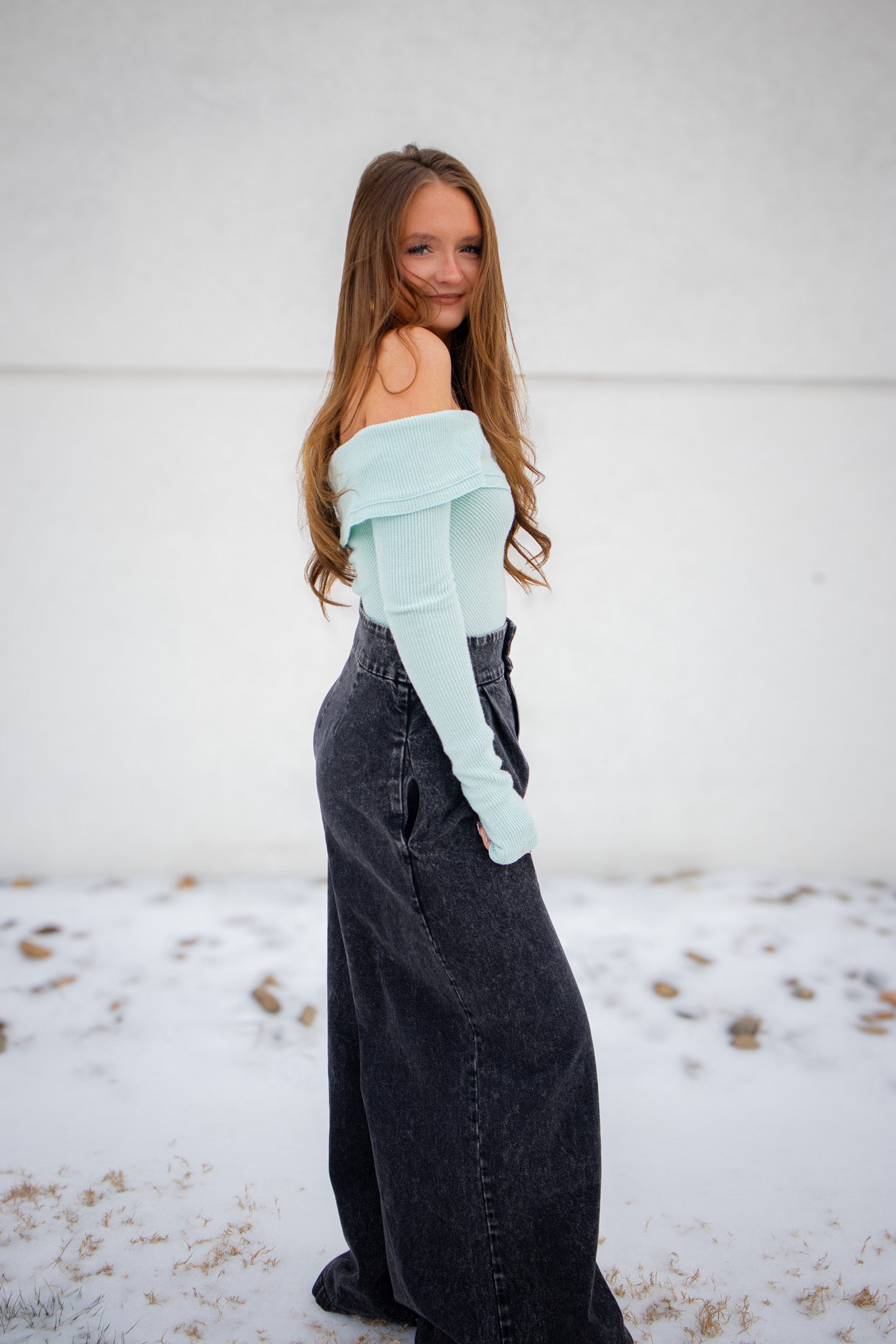 Person wearing a light blue off-shoulder top and dark jeans standing in a snowy landscape.