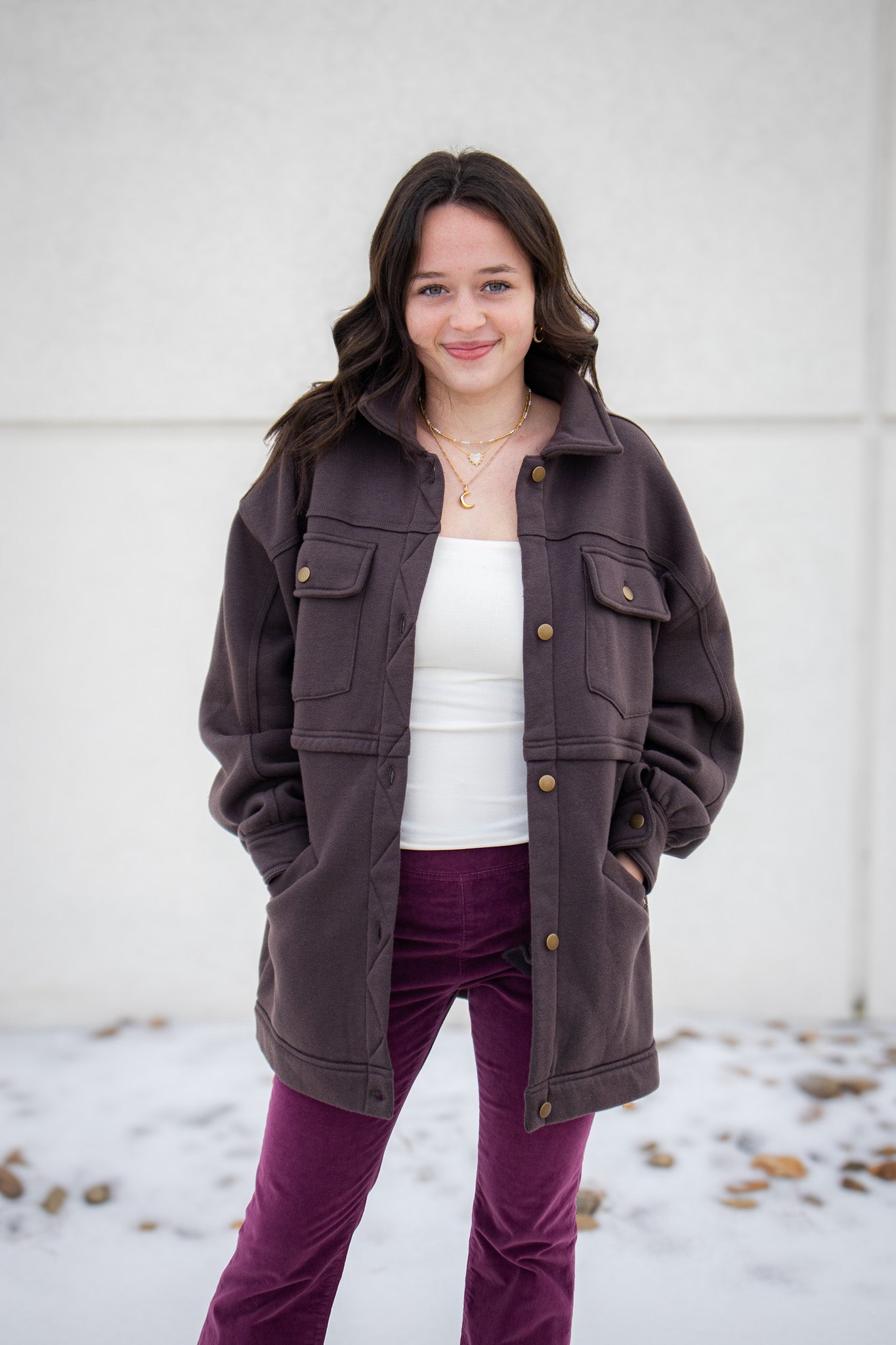 Woman wearing a brown jacket, white shirt, and purple pants standing against a light-colored wall.