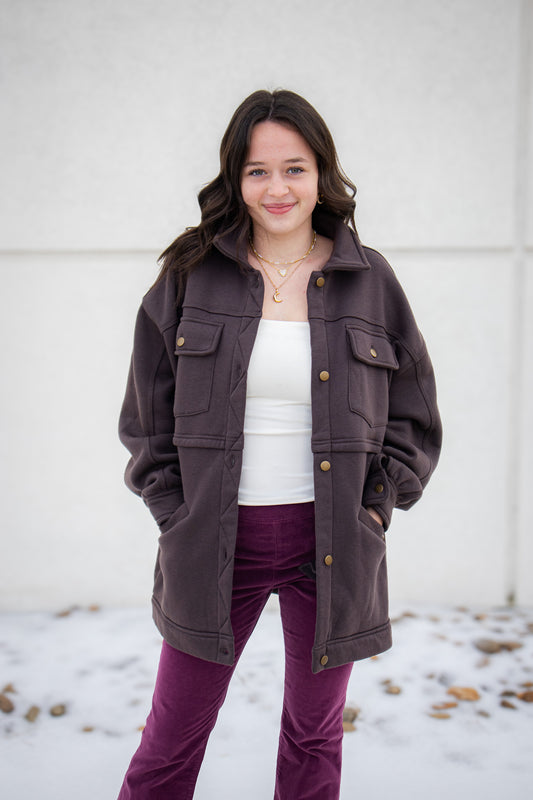 Woman wearing a brown jacket, white shirt, and purple pants standing against a light-colored wall.