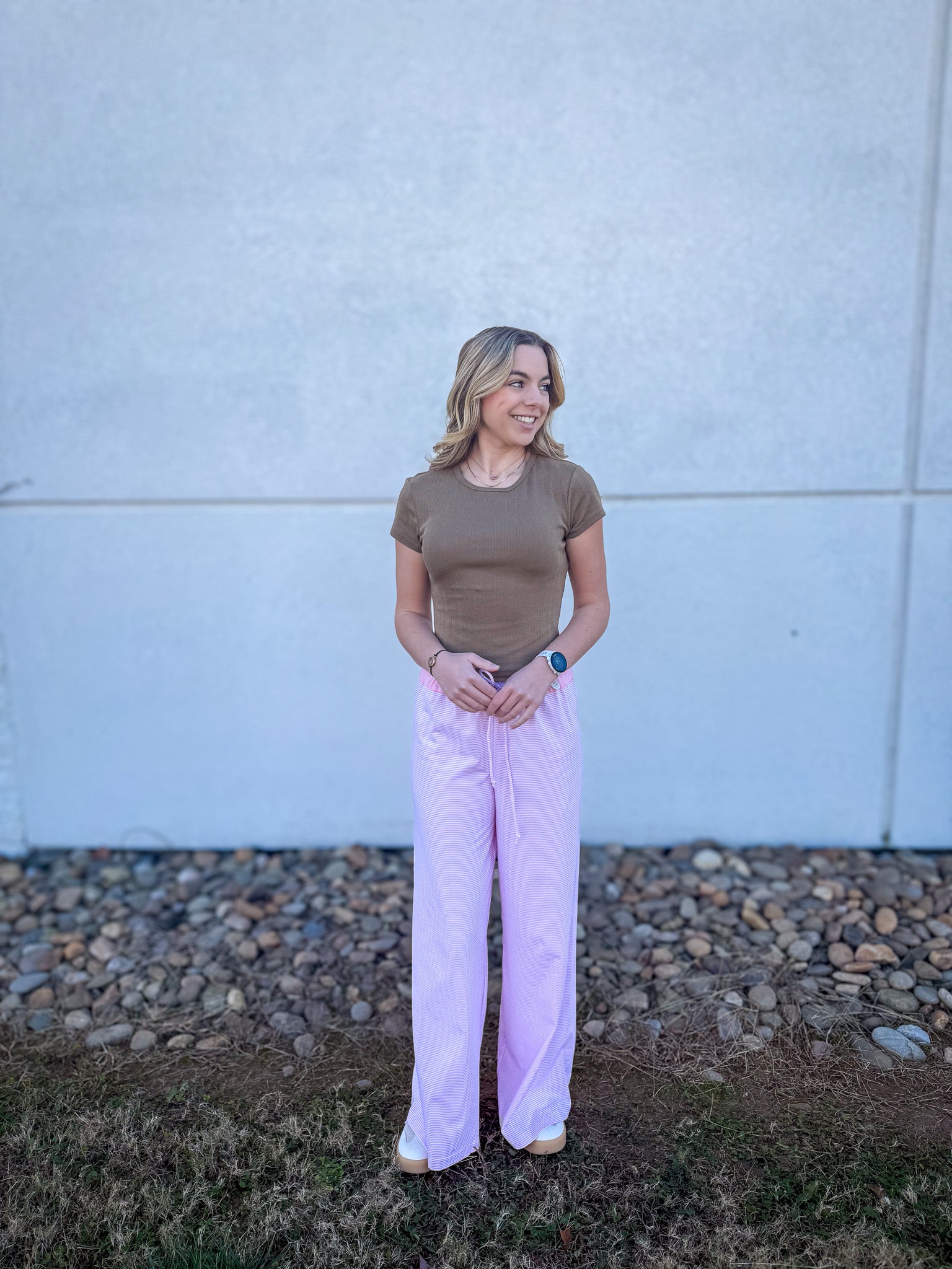 Woman wearing a brown top and pink pants standing against a light gray wall.