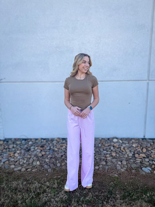 Woman wearing a brown top and pink pants standing against a light gray wall.