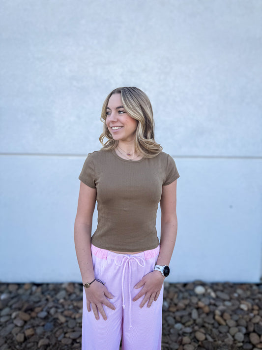 Woman wearing a brown t-shirt and pink pants standing against a light blue wall.