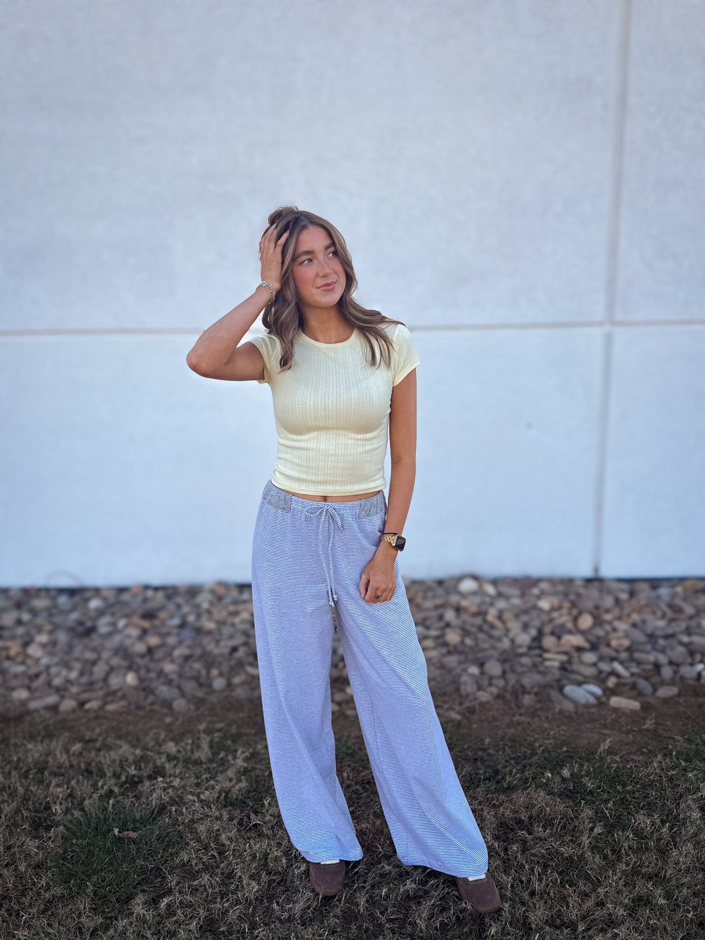 Woman wearing a yellow top and light blue pants standing against a white wall.