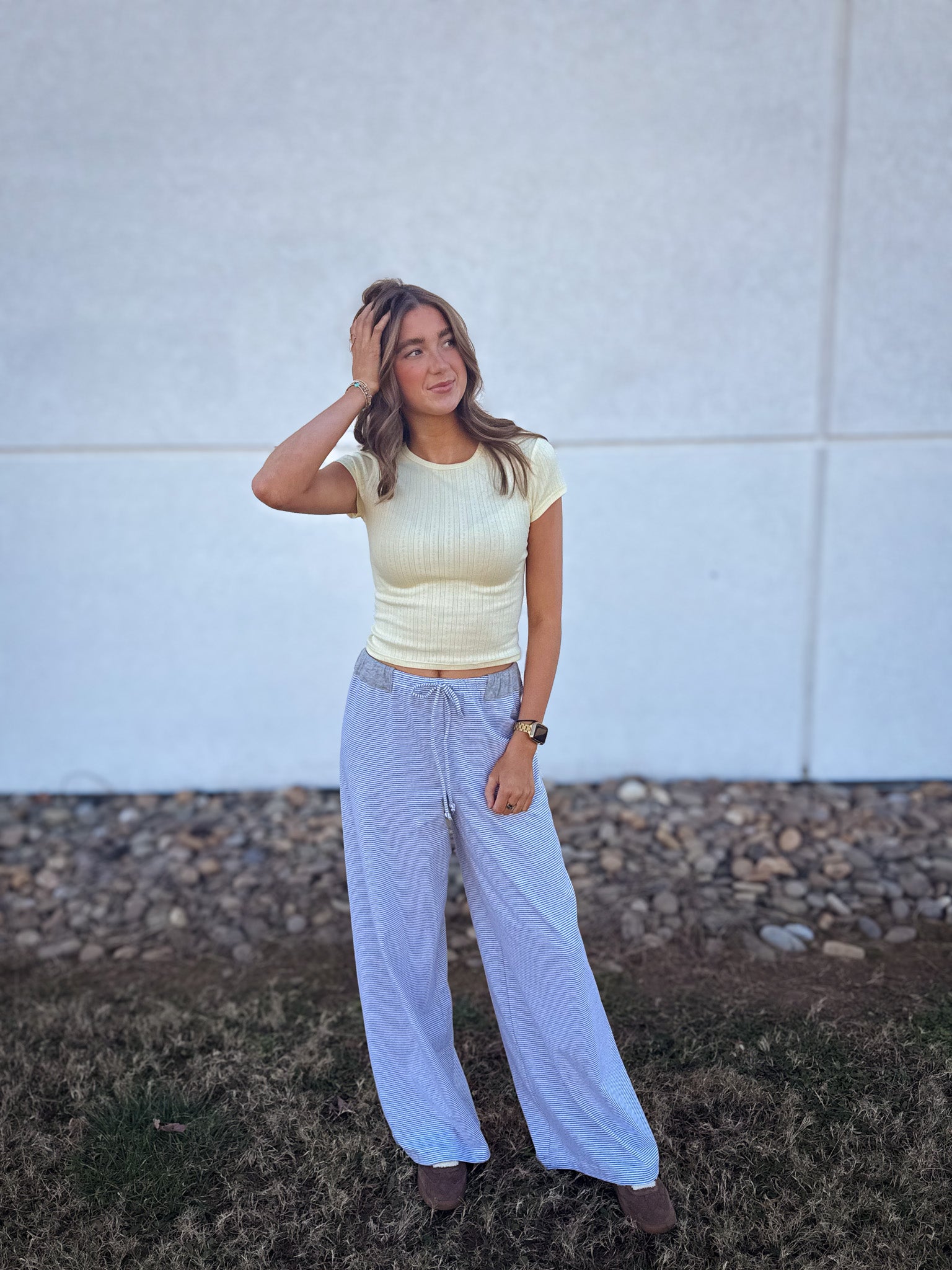 Woman wearing a yellow top and light blue pants standing against a white wall.