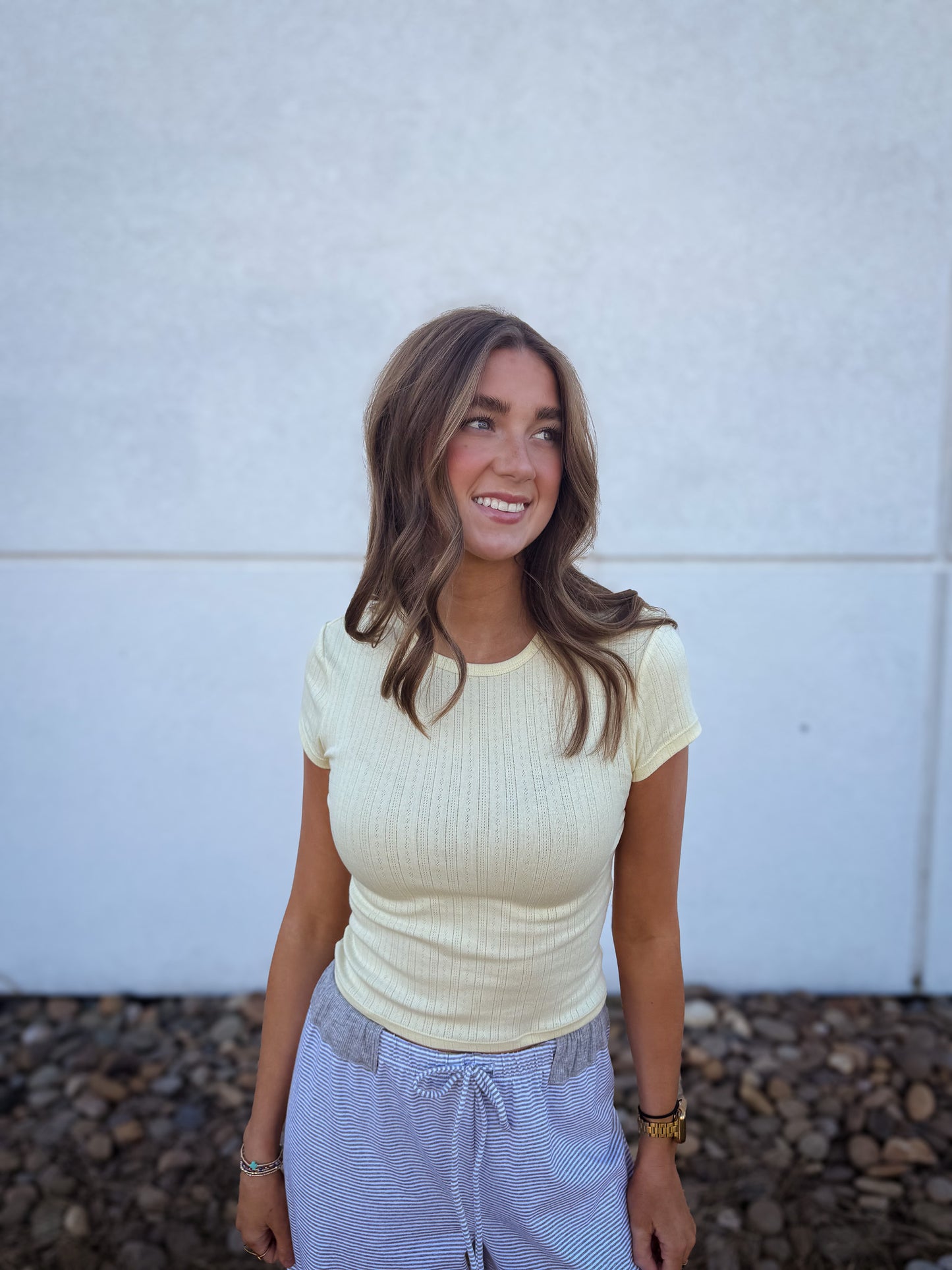 Woman wearing a light yellow ribbed top and gray pants against a white wall.