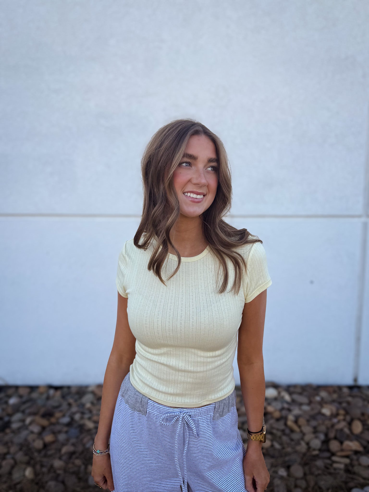 Woman wearing a light yellow ribbed top and gray pants against a white wall.