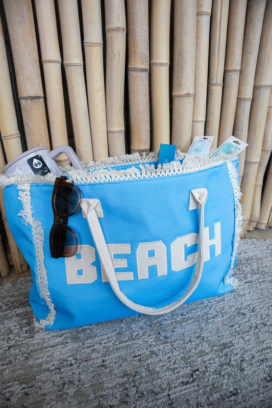 Blue beach bag with 'BEACH' text and sunglasses on a textured surface with bamboo background