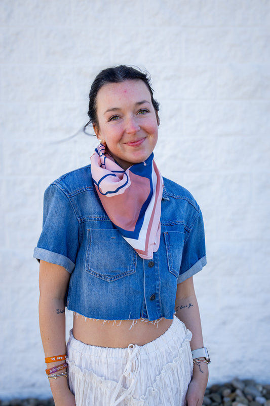 Woman wearing a denim jacket and white pants with a colorful scarf, standing against a white wall.