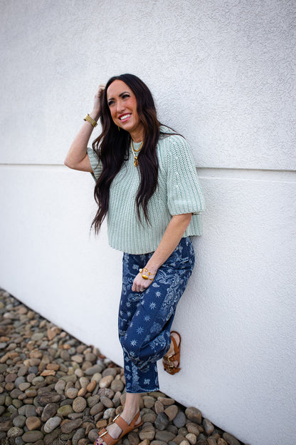 Woman wearing a light green sweater and patterned barrel jeans against a white wall.