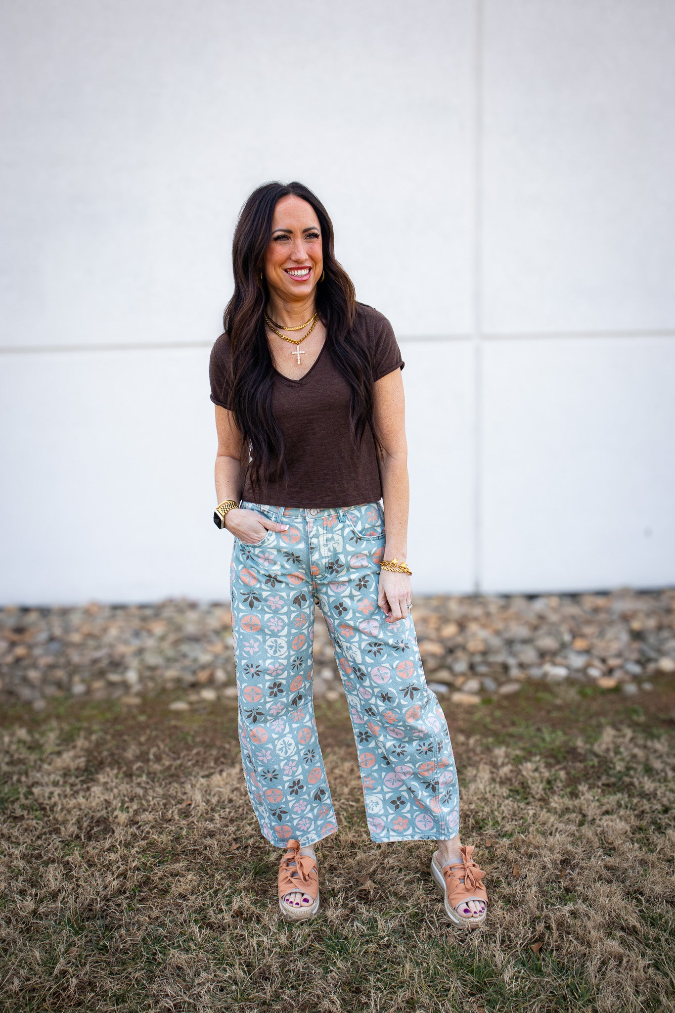 Woman wearing a brown tee and floral barrel leg jeans standing in front of a white wall.