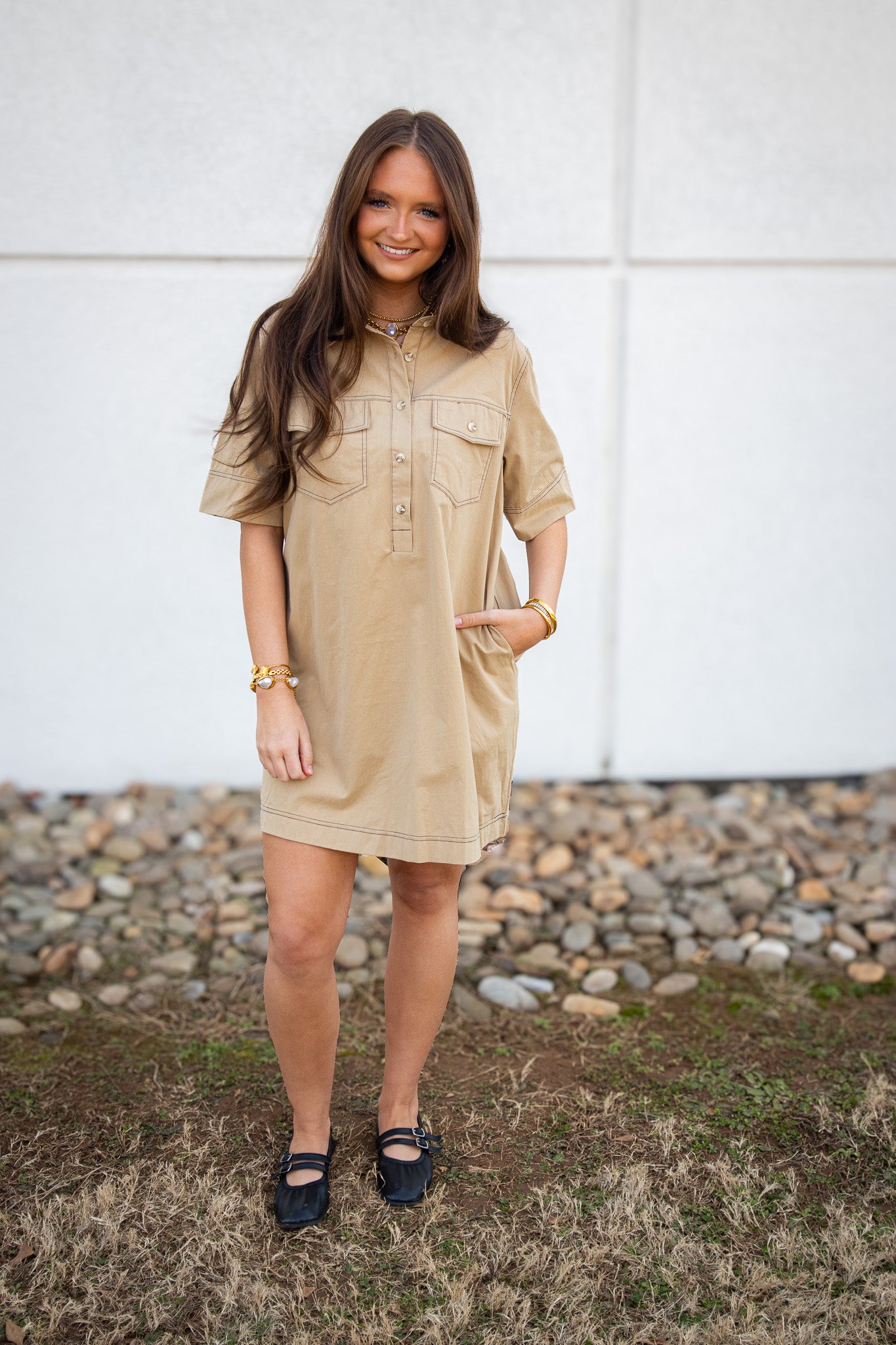 Woman wearing a beige dress standing outdoors with a white wall and stones in the background