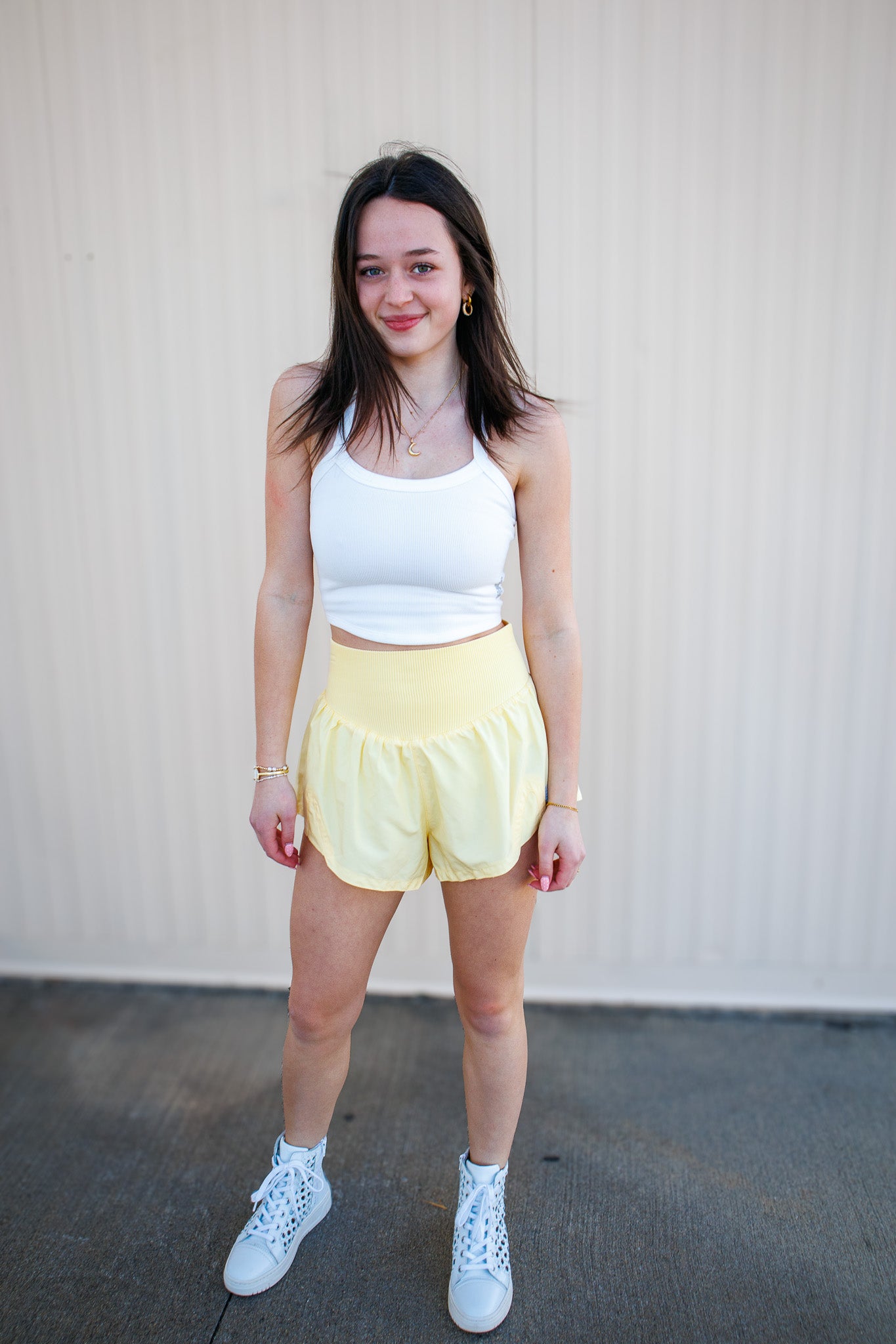 Woman wearing a white tank top and yellow shorts standing against a plain background