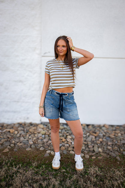 Woman wearing a striped t-shirt and denim shorts standing against a white wall.