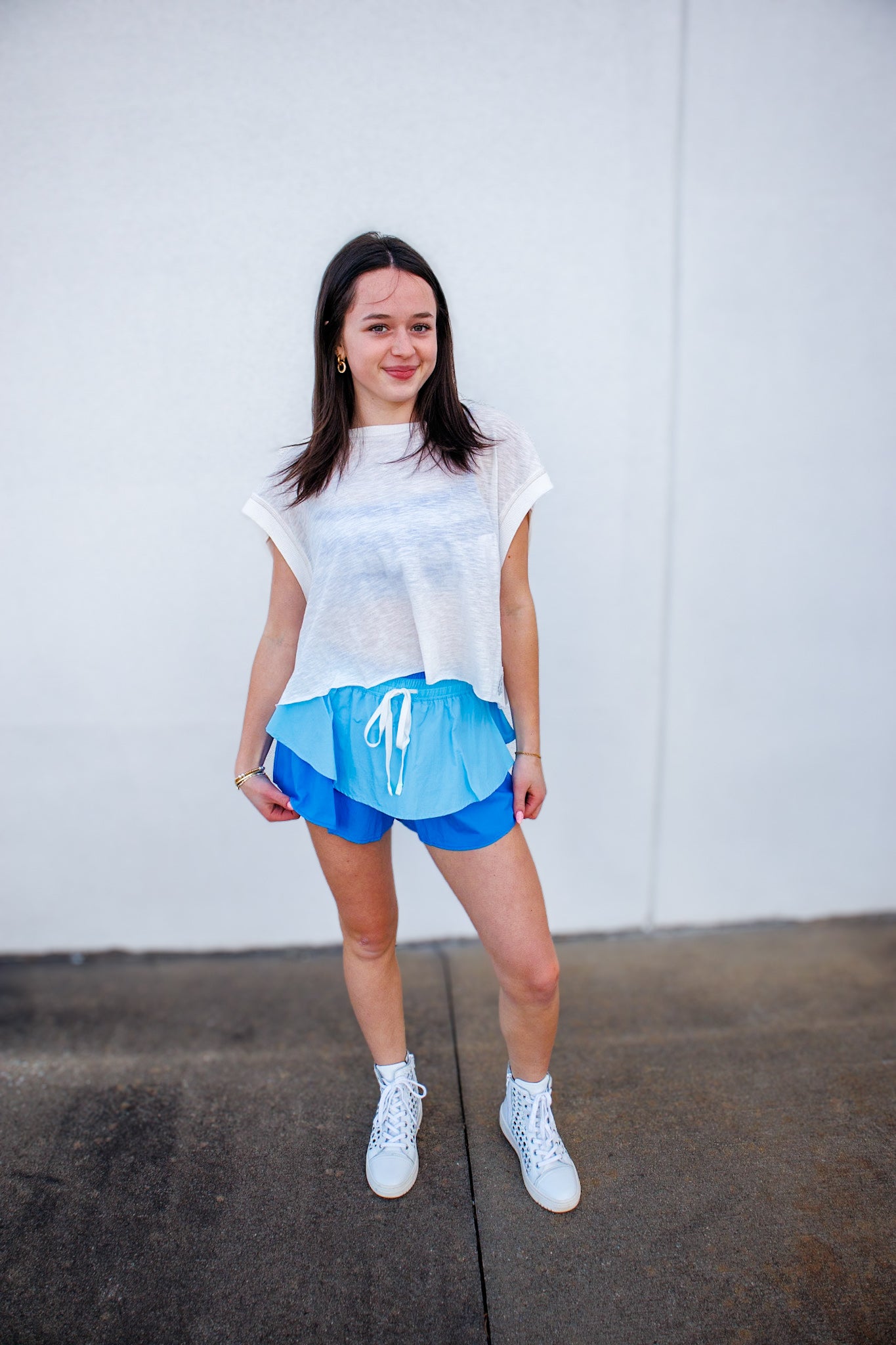 Woman wearing a white t-shirt and blue shorts standing against a plain wall.