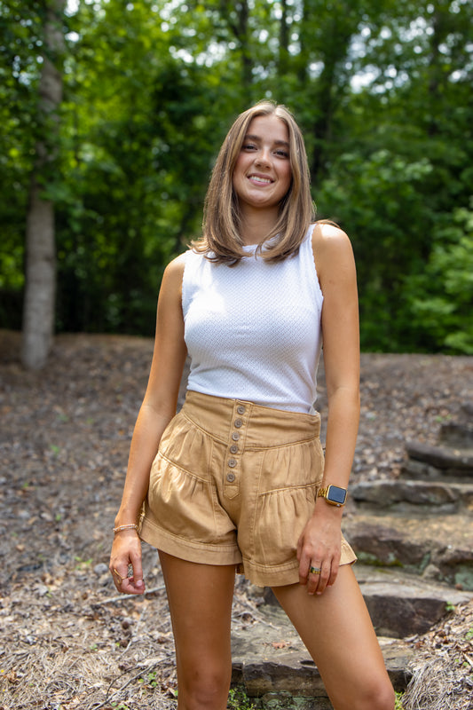 Woman in a white tank top and brown shorts standing outdoors with greenery in the background