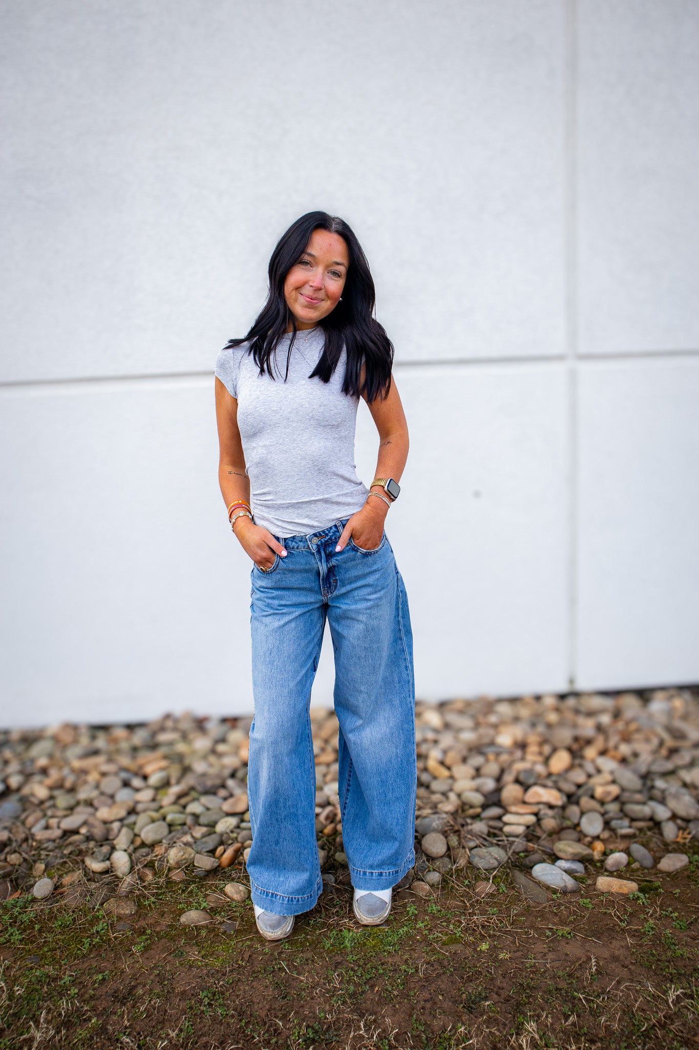 Woman wearing a gray t-shirt and blue jeans standing against a white wall with a stone ground.