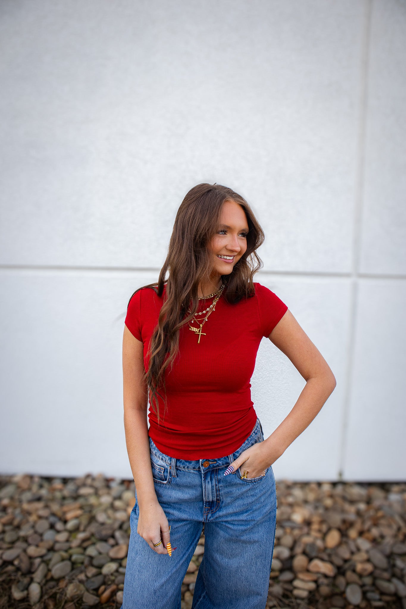 Woman wearing a red top and blue jeans standing against a white wall.