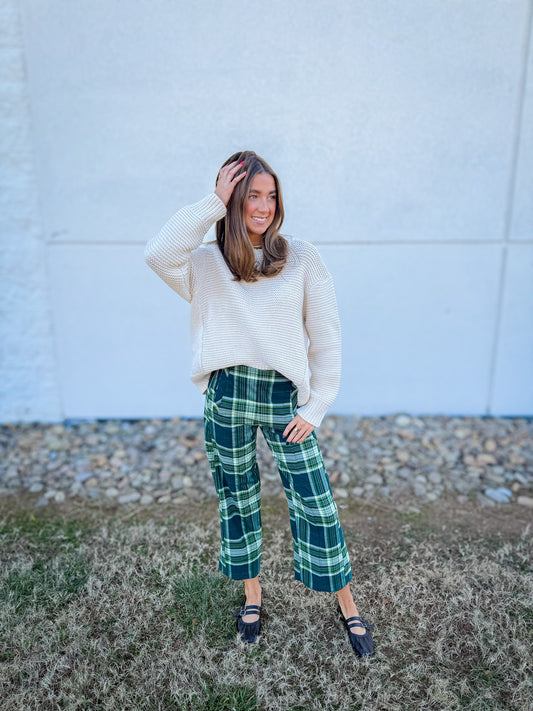Woman wearing a cream sweater and green plaid pants with black ballet flats standing against a light-colored wall.