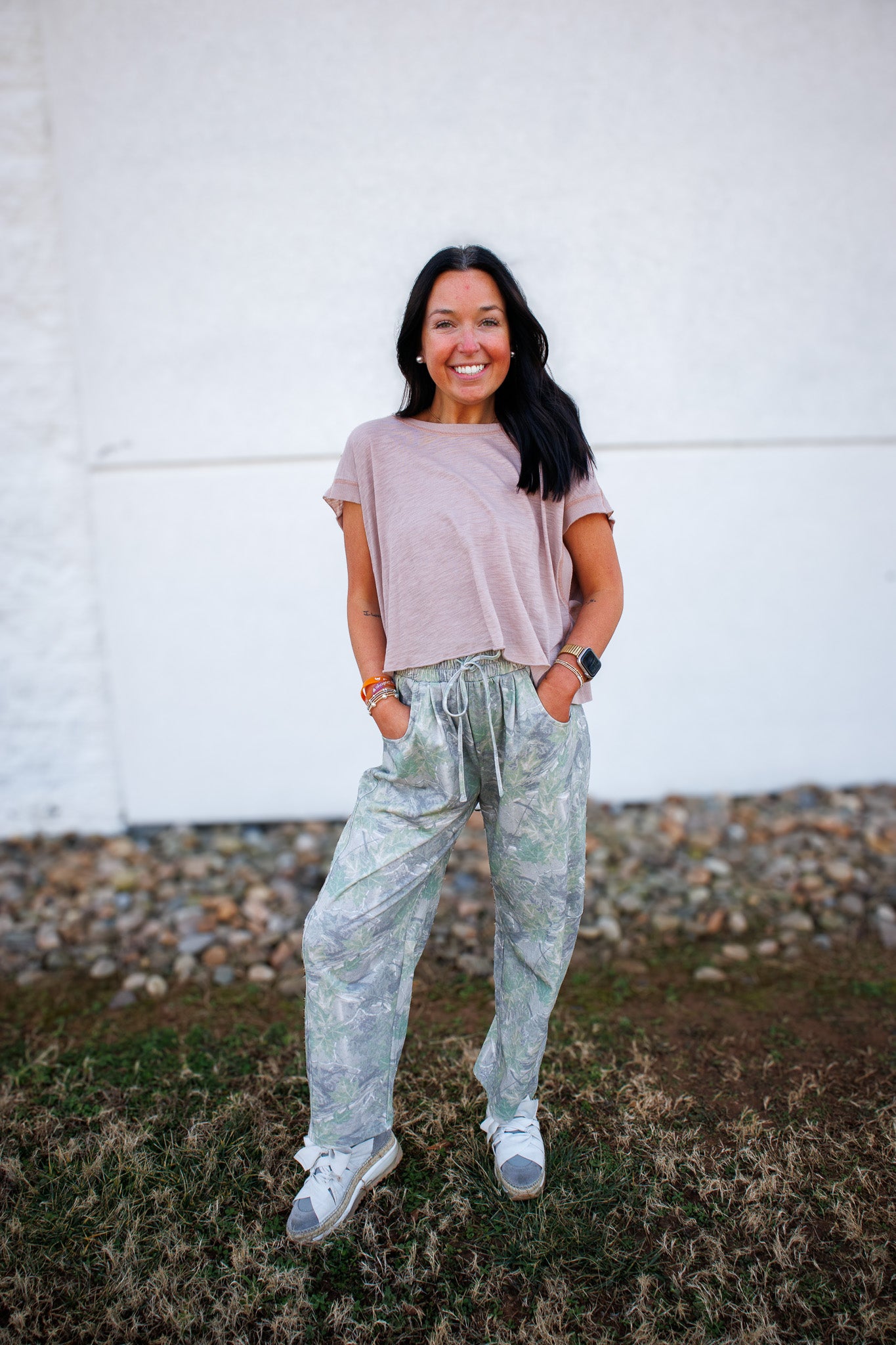 Woman wearing a loose-fitting t-shirt and camo pants standing against a white wall.