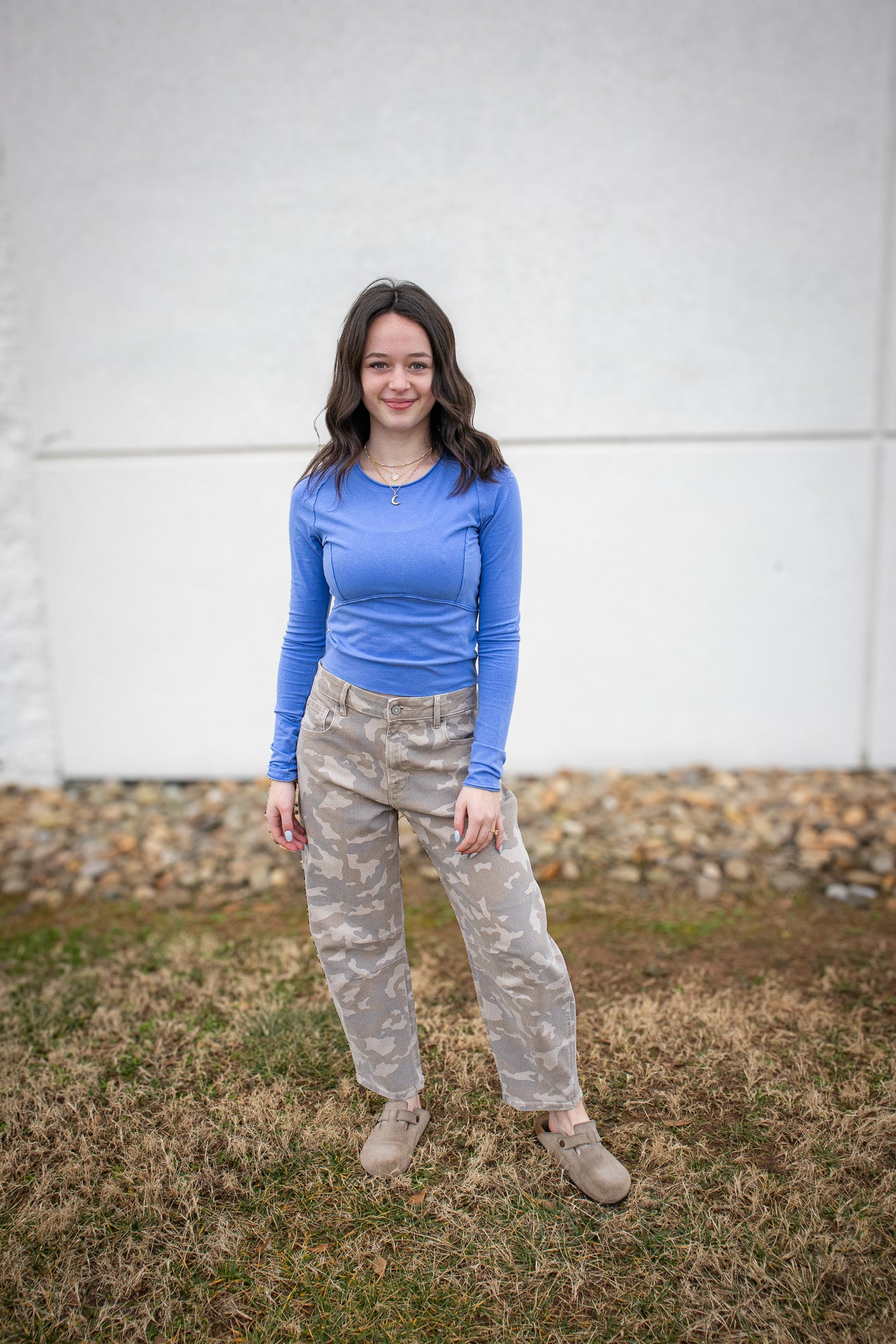 Person wearing a blue long-sleeve shirt and camouflage pants standing in front of a white wall.