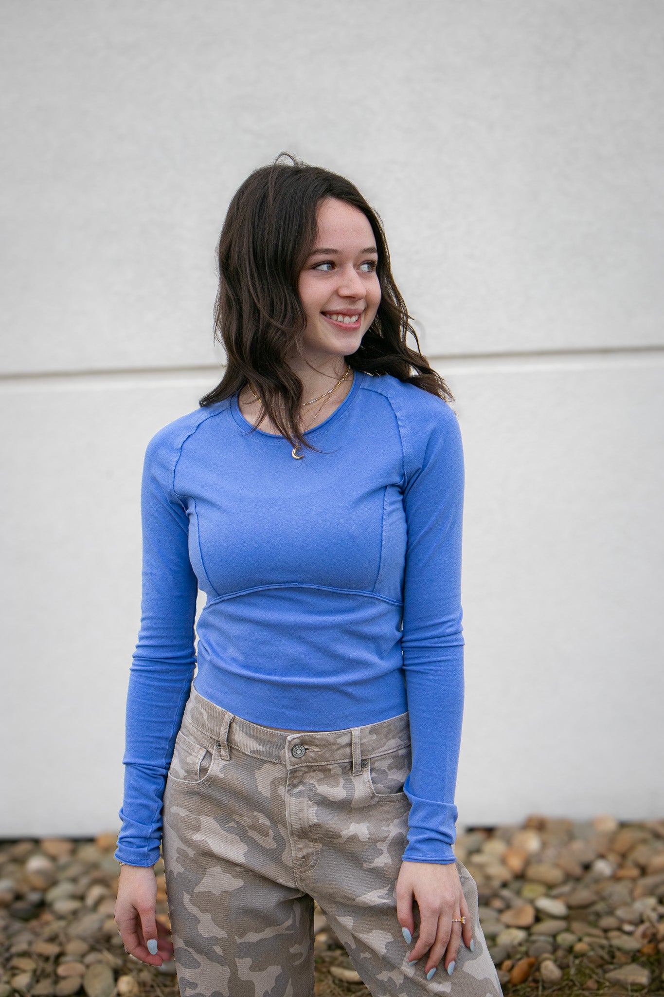 Woman wearing a blue long-sleeve top and camouflage pants standing against a light-colored wall.