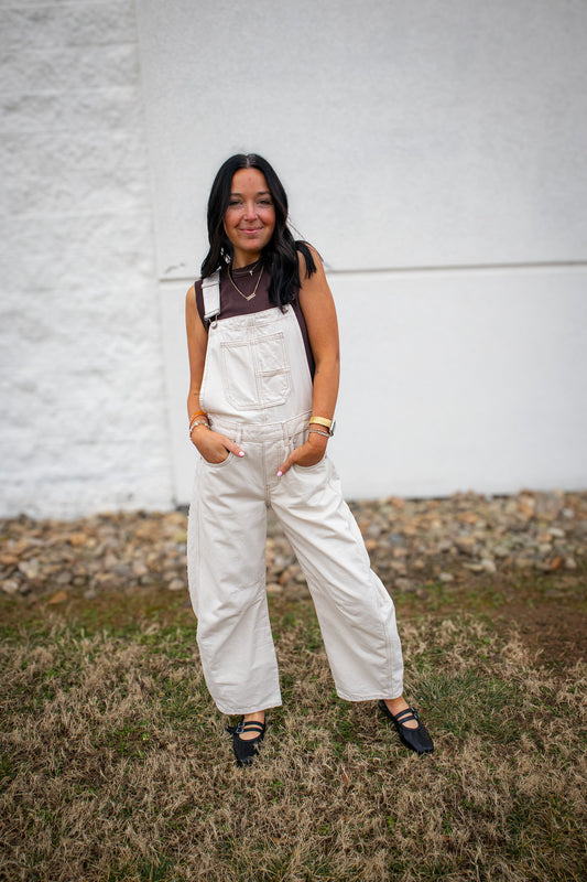 Woman wearing white overalls standing against a white wall.