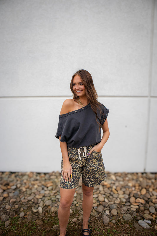 Woman wearing a black off-shoulder top and patterned shorts standing on a rocky ground with a light gray wall in the background.