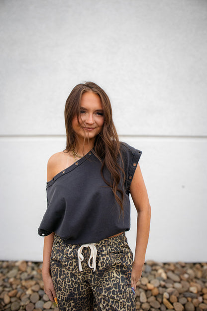 Woman wearing a dark gray off-shoulder top and leopard print pants against a white wall.