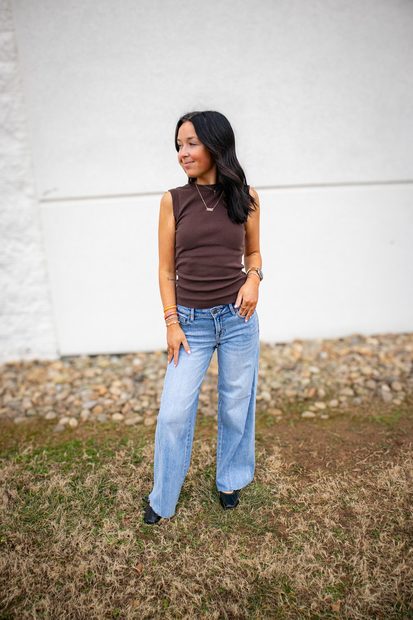 Woman wearing a brown sleeveless top and light blue jeans standing against a white wall.