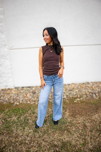 Woman wearing a brown sleeveless top and light blue jeans standing against a white wall.