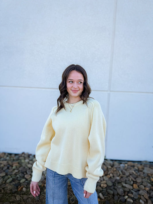 Woman wearing a light yellow sweater and blue jeans standing against a white wall.